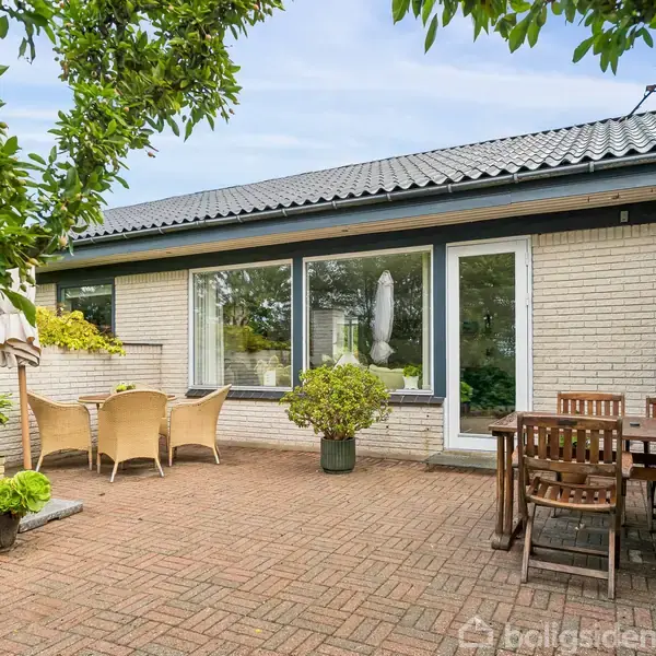 Terrasse med møbleret spisebord og stole på brolagt grund. En plantepotte står centralt blandt andre planter. Knyttet til en lys murstensbygning, omgivet af træer og grønt.