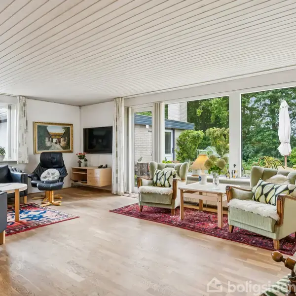 Stue med lyse træmøbler og grønne stole placeret på et rødt tæppe. Store vinduer i baggrunden viser en have. Moderne lamper hænger i loftet.
