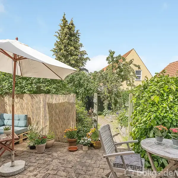 En lille, hyggelig terrasse med møbler inkluderer en parasol og flere planter i krukker. Omkranset af hække og træer i et boligområde med røde tegltage og en blå himmel.