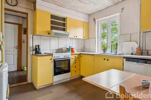 Et køkken med gule skabe og hvide fliser. En ovn og opvaskemaskine står under skabene, mens et bord med en krukke ses i forgrunden ved siden af et vindue.