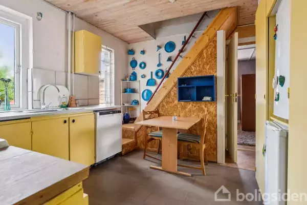 Køkken med gule skabe og bordplader, opvaskemaskine til venstre. Blå gryder og pander hænger på væggen ved trappen. Et lille bord med to stole står i hjørnet.