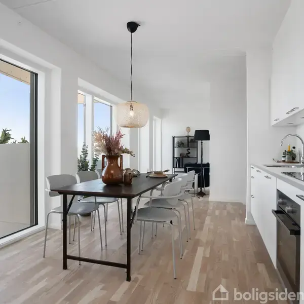 Et langt, moderne køkken med hvide skabe og trælaminatgulv. Et mørkt spisebord med stole står midt i rummet. Store vinduer giver udsigt til en balkon og natur.