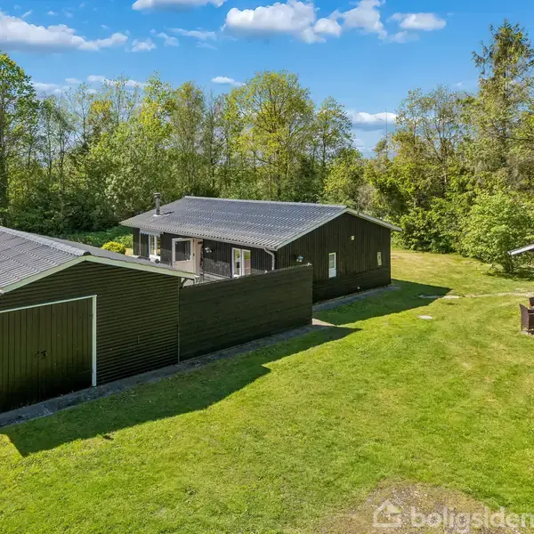 Et sort, enfamiliehus står i en frodig have med en garage ved siden af. Omgivet af grønne træer under en blå himmel med lette skyer.