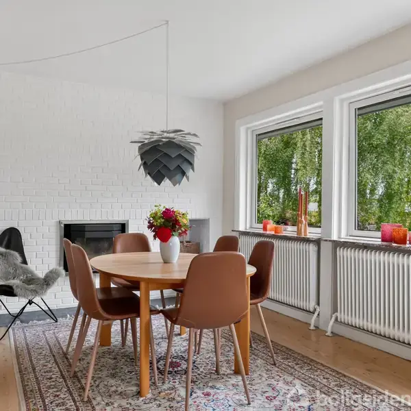 Et rundt spisebord med stilfulde brune stole står i et lyst rum. Bordet er pyntet med en blomsterbuket. Baggrunden har to store vinduer med udsigt til træer.