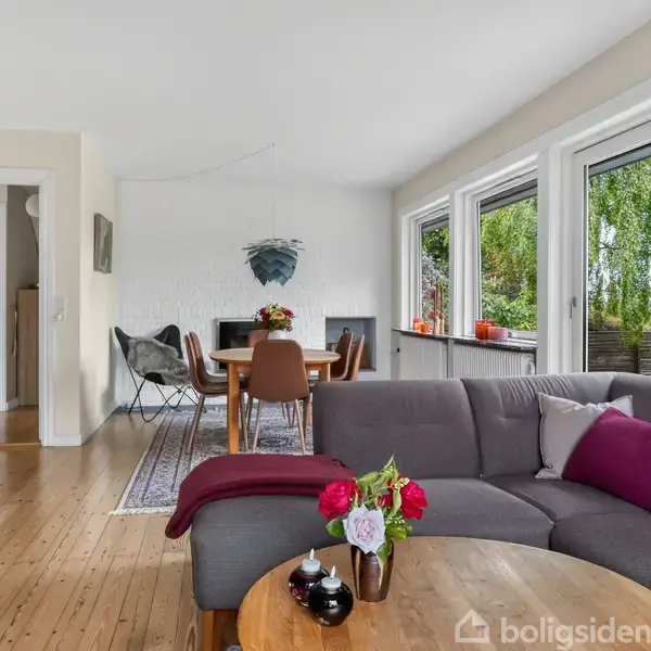 Grå sofa med puder står foran runde sofaborde med blomster. Spisestue i baggrunden med sort bord og stole. Store vinduer til venstre med udsigt til grønne træer.