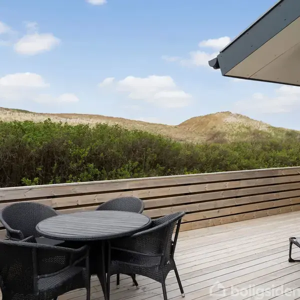 Rundt havebord med fire stole står på en træterrasse ved et sommerhus. I baggrunden ses sandklitter og buskadser under en blå himmel med lette skyer.