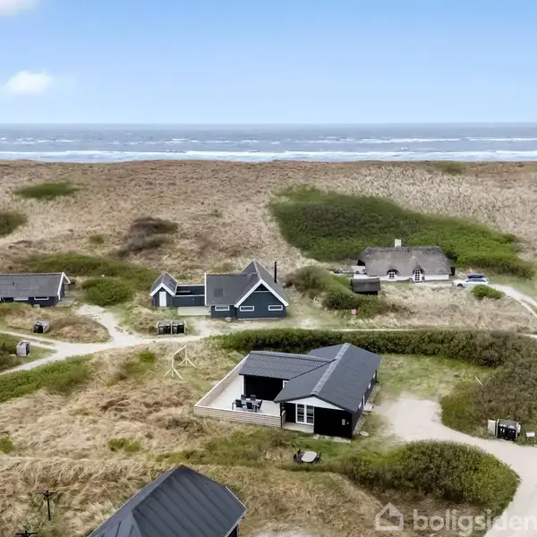 Flere sommerhuse ligger spredt blandt grønne klitter nær en kystlinje. Stier forbinder bygningerne, mens havet kan ses i baggrunden under en let skyet himmel.