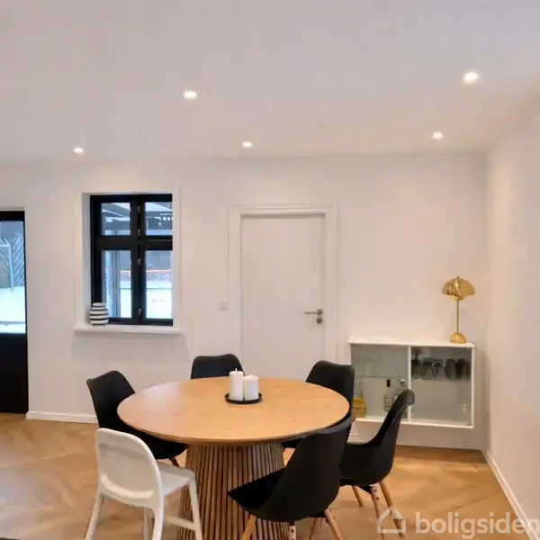 Rundt spisebord med fem sorte stole og én hvid stol står i et moderne, lyst køkken. En gulvlampe er placeret på et hvidt skab med glaslåger mod væggen.