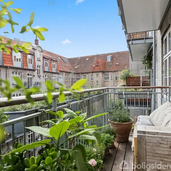 Balkon omkranset af grønne planter og træmøbler. Udsigten inkluderer nabobygningers stenfacader med røde tegltage. En lys, klar himmel over de tætte bymæssige omgivelser giver hyggelig stemning.
