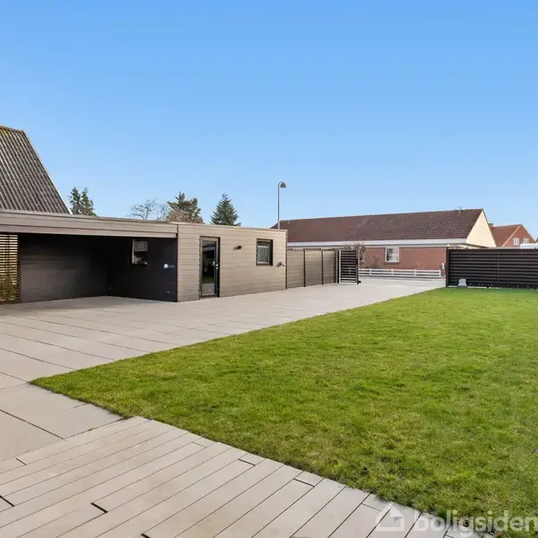 Carport med fladt tag står på flisebelagt terrasse ved stor græsplæne i en boligkvarter. Omkringliggende hegn og naboers huse ses i baggrunden under en klar blå himmel.