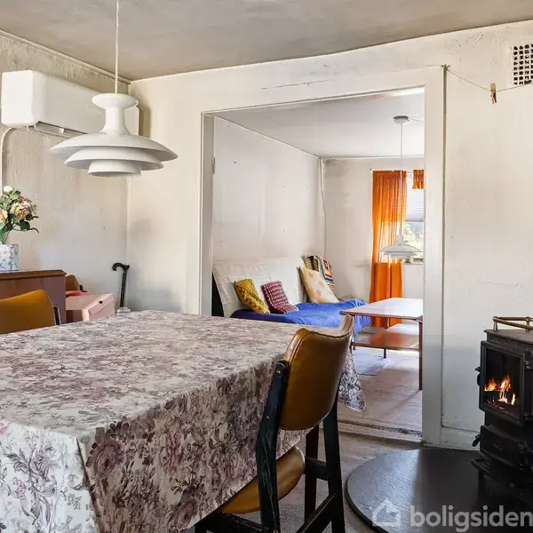 Et spisebord med blomstret dug står i et rum med hvide vægge; en brændeovn til venstre og blomsterdekoration på et skab. I baggrunden ses en stue med sofa.