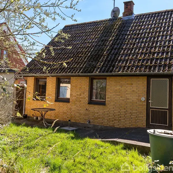 Et gult murstenshus med skråt tag står stille i en solrig haveomgivelse. En grøn affaldsbeholder og et lille rundt bord er placeret på den grønne græsplæne.