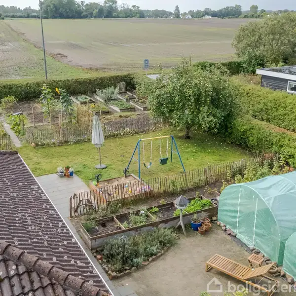 Et lille haveområde med en gynge og grønne planter omgivet af hegn. To drivhuse står ved siden af en terrasse med havemøbler. Åbne marker breder sig i baggrunden.