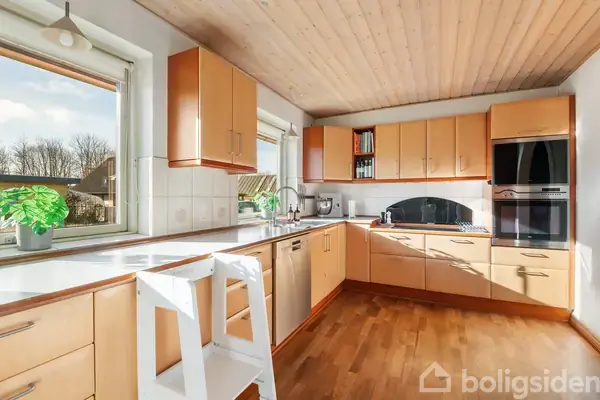 Et lyst køkken med trægulv og træloft. Køkkenskabe er lysebrune, og der er en indbygget ovn. En lille trappestige står foran en stor bordplade med grøn plante.