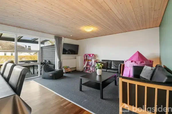 Stue med sort sofa og sofabord, børnelegetøj og fjernsyn på væggen. Ved siden af står en pink legetelt. Rummet har et trægulv og leder ud til en overdækket terrasse.