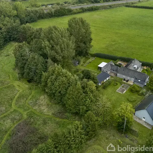 Grunden er indhegnet med blå linje og indeholder tætte træer og en bygning. Den ligger i en grøn, landlig omgivelser nær en by med huse i baggrunden.