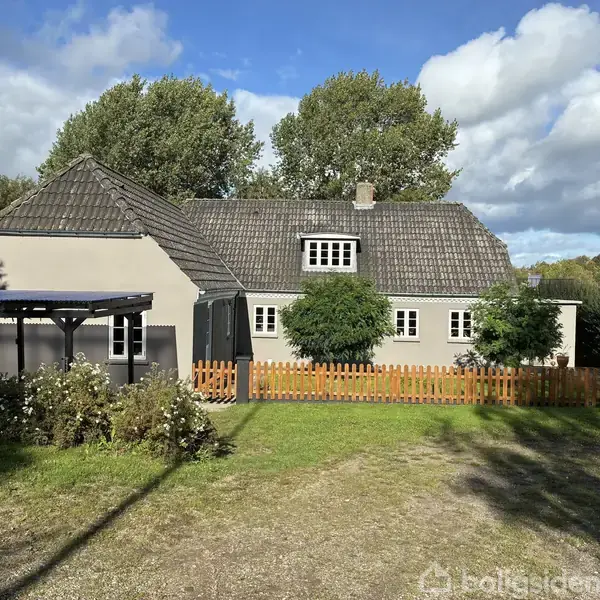 Et charmerende gråmalet hus med mørkt tag står stille omgivet af blomster og træer. En lille træhavelåge og en carport til venstre skaber en idyllisk scene.