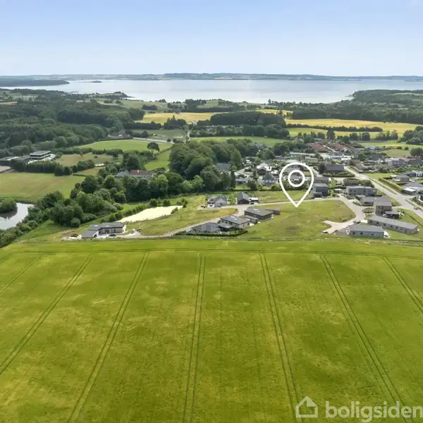 En ejendom er markeret med en stedsmarkør i en grøn landlig landsby. Omkringliggende områder består af marker, skove og en stor sø i baggrunden.