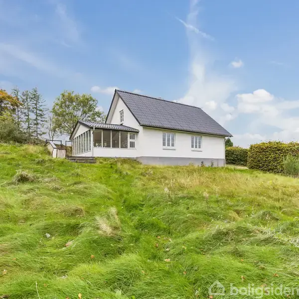 Hvidt hus med sort tag står stille omgivet af en grøn, let kuperet græsplæne. Baggrunden har træer og blå himmel med få skyer.