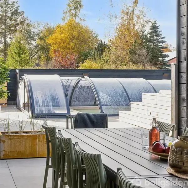 En udendørs træterrasse med et langt bord og stole står i forgrunden. I baggrunden ses en overdækket swimmingpool omgivet af frodige træer med efterårsfarver.