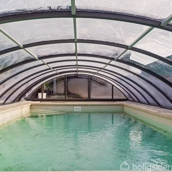 En indendørs swimmingpool er dækket af en gennemsigtig kuppel. Vandet er roligt, og kuplen beskytter poolen mod omgivelserne. I baggrunden ses en plante i en potte.