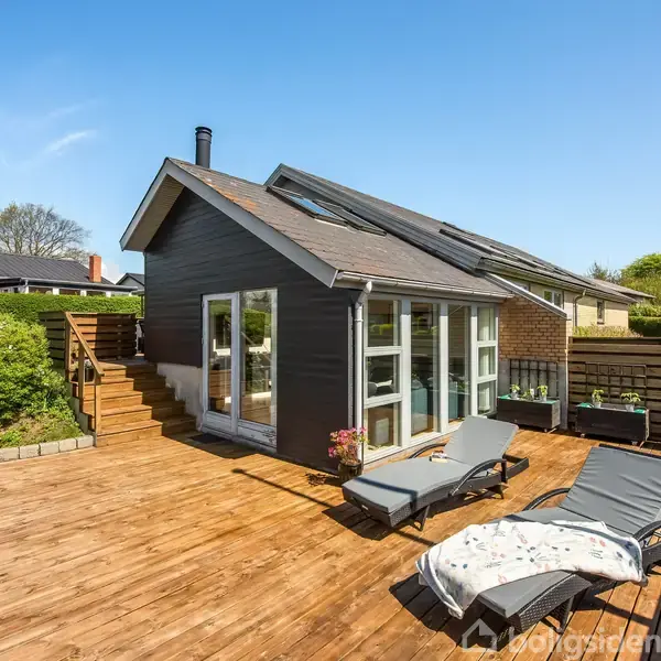 Et moderne træhus med store glasvinduer står på en solrig træterrasse. To liggestole med tæpper er placeret foran huset, omgivet af grønne hække og en klar blå himmel.
