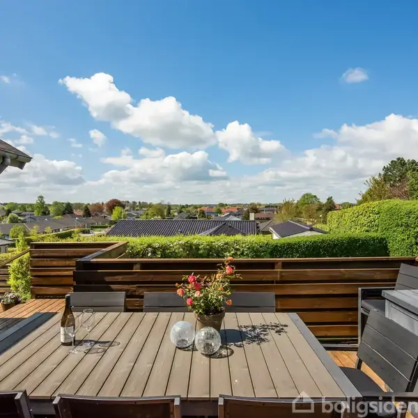 Terrassebord med stole står udendørs på en terrasse. Bordet har en vase med blomster og dekorationer. I baggrunden ses udsigt over hække og hustage under en blå himmel med skyer.