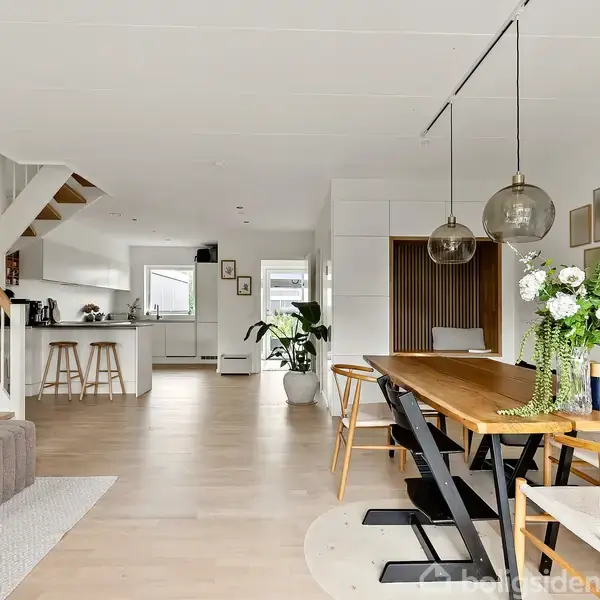 Spisebordet i træ står i en moderne stue med en åben planløsning, hvor det er dækket med blomster. I baggrunden ses et lyst køkken og en trappe til overetagen.