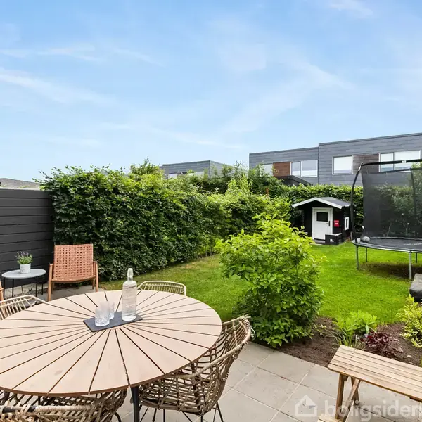 Rundt træbord med stole står på flisebelagt terrasse. I haven ses en trampolin, et lille legehus og grønne buske omgiver området. Skyfri himmel i baggrunden.