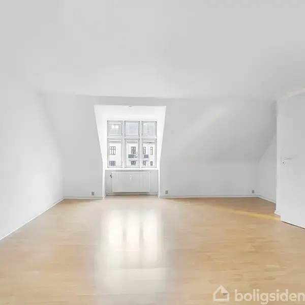 Et tomt rum har lyst trægulv og hvidmalede vægge. Sollys strømmer ind fra et vindue, der dominerer bagvæggen. Rummet fremstår rummeligt og minimalistisk.