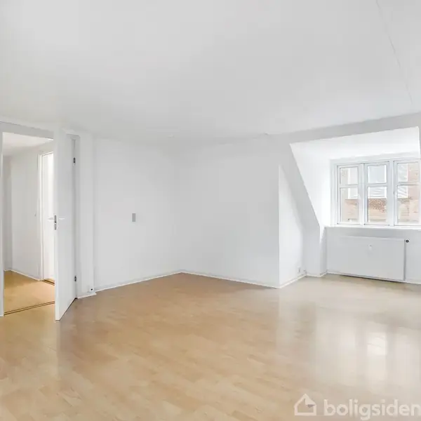 Et lyst rum med trægulv og skrå lofter. En åben dør fører til en tilstødende gang. Et vindue med udsigt tilbygninger giver naturligt lys.