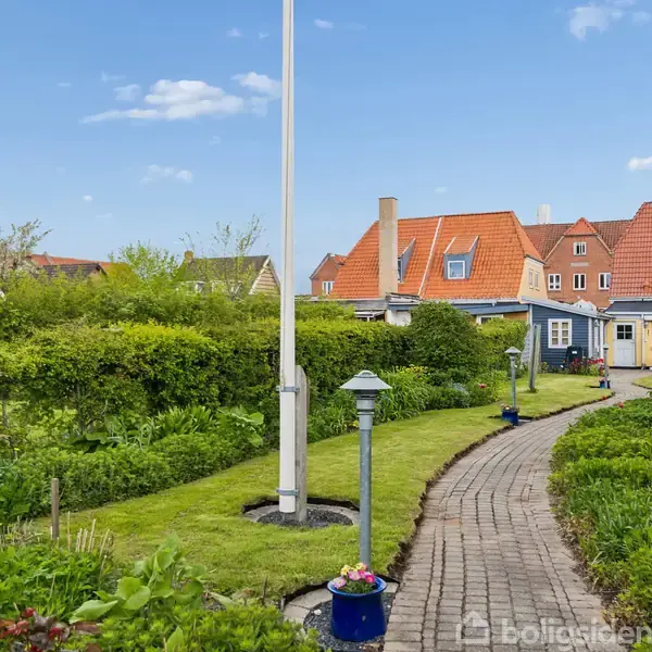 En snoet sti leder gennem en velplejet have med blomster og buske. I baggrunden ses et hus med røde tage og flere kviste under en blå himmel.