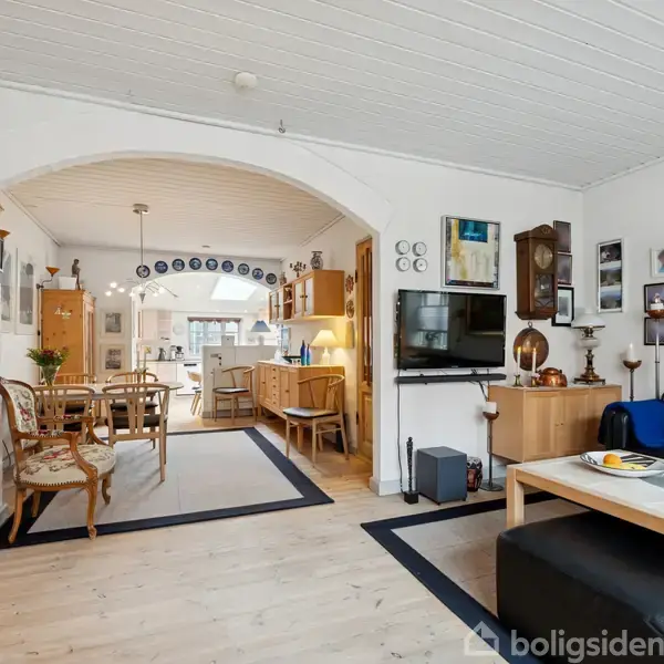 Stue med sofa og sofabord til højre, TV på væggen, træmøbler og billeder. Spisestue med bord og stole ses gennem en bueåbning. Lyst og hyggeligt indrettet.