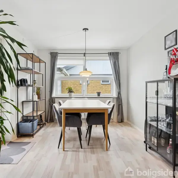 Et spisebord står i et lyst rum med grå gardiner, omgivet af en reol til venstre og en vitrineskab til højre. En stor plante står ved siden af en sofa.