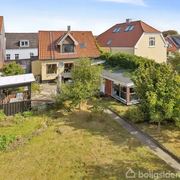 Et hyggeligt parcelhus med rødt tag står i en have med græs, træer og en flisebelagt sti. Et skur ses til venstre ved siden af en lille terrasse.