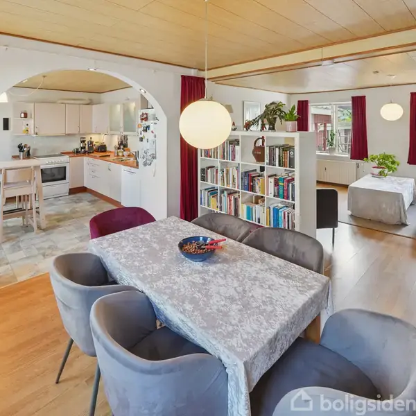 Spisebord dækket med gråt dug og skål, omringet af grå stole. I baggrunden ses en hvid bogreol, et lyst køkken og en stue med røde gardiner og store vinduer.