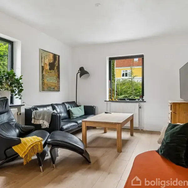 Stue med sorte læderstole, en sofa og et sofabord. En lampe står ved siden af sofaen. Vinduerne viser udsigt til en have. En plante står på en vindueskarm.