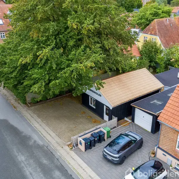 Hus med skråt tag omgivet af træer og andre bygninger. En sort bil er parkeret ved indkørslen på en brolagt gårdsplads. Vej løber langs ejendommen med fortov.