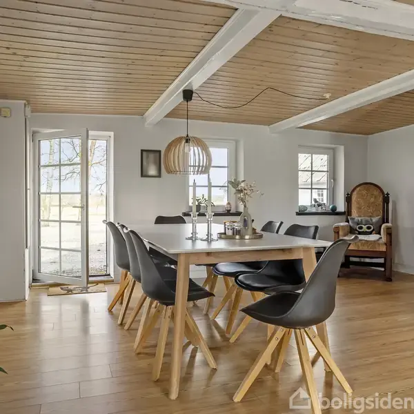 Spisebord omgivet af seks sorte stole står på trægulv under træloft med hvide bjælker. En moderne lampe hænger over bordet, og der er vinduer og dør i baggrunden.