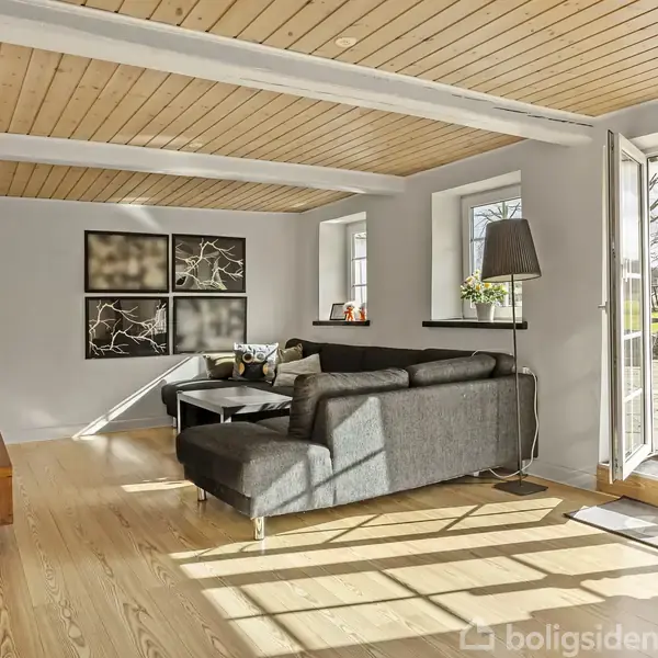 Grå hjørnesofa står i en stue med trægulv og træloft. Sofaen vender mod et sofabord foran vægkunst. Glasdøre åbner til en terrasse, der leder til en græsplæne.