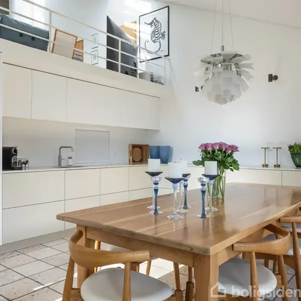 Et lyst køkken med hvidt inventar, hvor et træbord står centralt omgivet af stole. På bordet er der lysestager og blomster. En moderne loftlampe hænger ovenover.