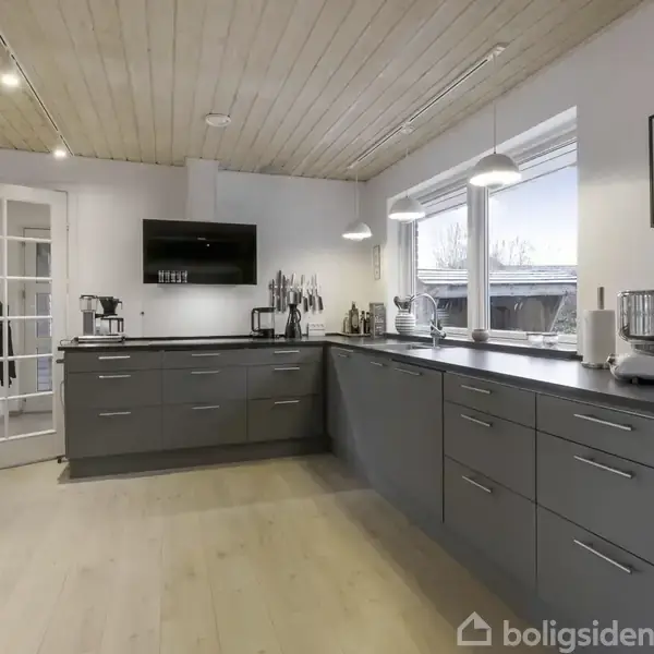 Moderne køkken udstyret med grå skabe og apparater; to store vinduer lader dagslys ind; lyse trægulve kontrasterer loftpaneler; en glasdør fører til en anden del af huset.