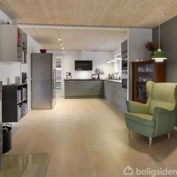 En moderne stue-køkken-kombination med en tændt brændeovn i venstre side, grøn lænestol til højre, og moderne køkken med grå skabe i baggrunden. Verdenskort hænger på væggen.