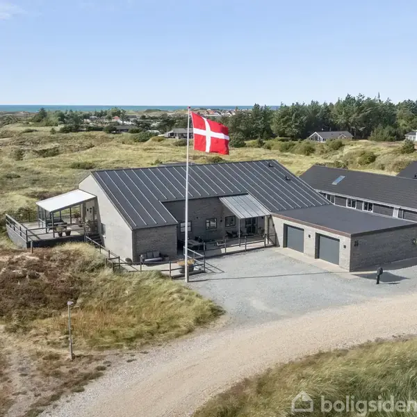 Et moderne hus med mørkt tag og flagstang med et dansk flag vejrer i vinden nær sandede klitter. Flere huse i baggrunden og havet i horisonten.