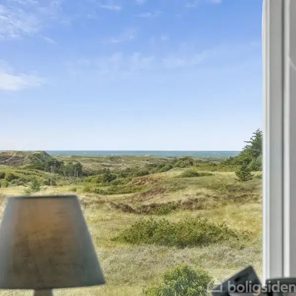 En lampe står foran et stort vindue med udsigt over et frodigt landskab med bakker og hav i horisonten. Himlen er klar og blå.