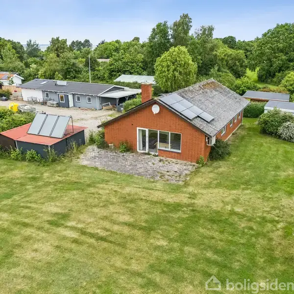 Et rødt hus med solpaneler på taget står på en stor, veltrimmet græsplæne. Ved siden af er en mindre bygning med solpaneler. Omkranset af tæt grøn vegetation.
