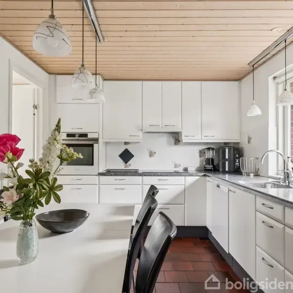 Et køkken med hvide skabe og apparater, der inkluderer en indbygget ovn. En hvid spisebord med sorte stole har en vase med blomster. Store vinduer slipper lys ind.