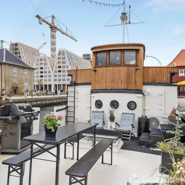 En husbåd på en kanal med siddemøbler og planter på dækket. I baggrunden ses moderne og traditionelle bygninger samt en kran.