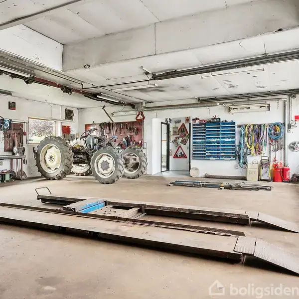 En værkstedshal med en traktor parkeret ved siden af arbejdsstationer. Vægge er dekoreret med værktøj og skilte. Rummet har store vinduer og porte.