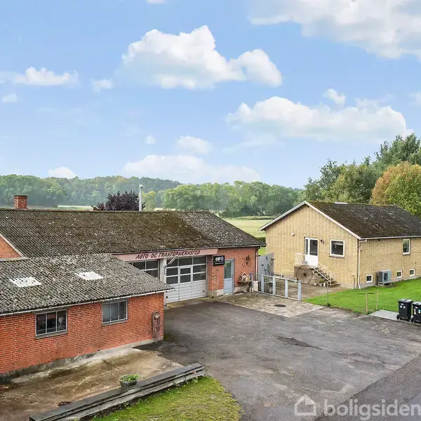 To bygninger, en rød murstensbygning med garageport og en gul villa, står i et landligt område med træer og grønne områder. Tekst på bygningen: "Aut. Elinstallatør".
